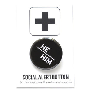 Round pinback button with black background which reads HE/HIM on an angle in white text, in a san serif font. On a white backing card, black plus sign at the top. Below reads: Social Alert Button, with tiny text beneath it which says, for common physical & psychological situations