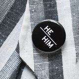 Round pinback button with black background which reads HE/HIM on an angle in white text, in a san serif font. Pinned on a white & gray textile blazer lapel