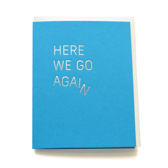 A bright medium blue greeting card with off white envelope, reads HERE WE GO AGAIN in a thin metallic silver foil, sans serif font. The word AGAIN starts to slide off, creating the image of the word coming apart and falling downward