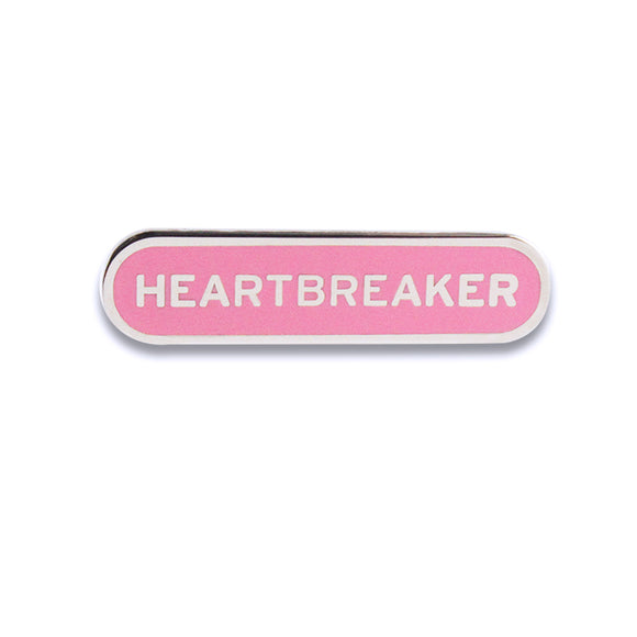 Capsule shaped enamel pin that says HEARTBREAKER.  Silver text and outline on a pink enamel background.