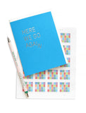 A bright medium blue greeting card with off white envelope, reads HERE WE GO AGAIN in a thin metallic silver foil, sans serif font. The word AGAIN starts to slide off, creating the image of the word coming apart and falling downward. Greeting card is sitting on a sheet of multicolor passage stamps with a pale pink pen with forest green text in French.