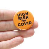 Orange HIGH RISK FOR COVID pinback button in an open palm for size reference.