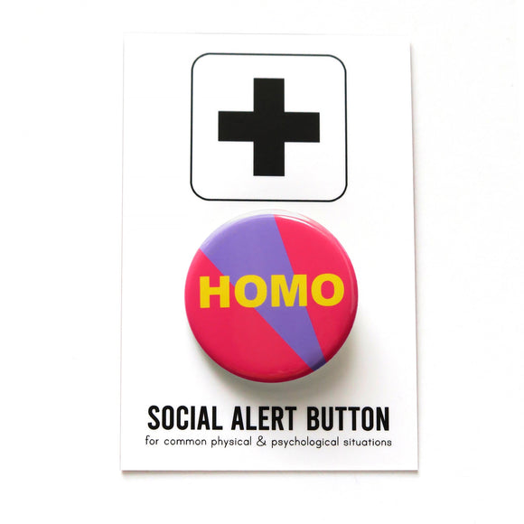Round pinback button with neon red-pin background with violet stripe which reads HOMO in bold yellow text. On a white backing card, black plus sign at the top. Below reads: Social Alert Button, with tiny text beneath it which says, for common physical & psychological situations