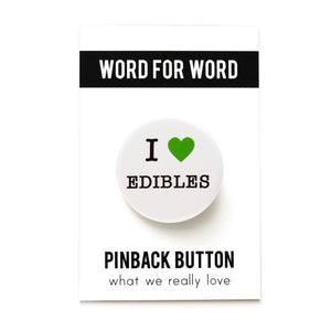 Round white button that reads I LOVE EDIBLES. Love is indicated with a green heart symbol, the remaining text is black. Button is on a Word For Word branded backing card.