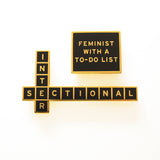 Crossword puzzle shaped enamel pin that says INTERSECTIONAL. And a rectangles shaped pin that says FEMINIST WITH A TO-DO LIST.