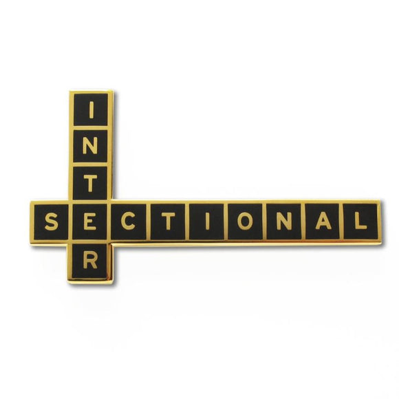 Crossword puzzle shaped enamel pin that says INTERSECTIONAL.  Gold text and outline on ad black enamel background.