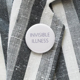 Round pinback button that reads INVISIBLE ILLNESS. Thin blue text on a white background. Button is pinned on white & gray woven blazer lapel.
