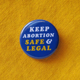 A round bright blue button that reads Keep Abortion Safe & Legal in retro yellow & white text.  Badge is pinned to a mustard yellow cotton shirt.