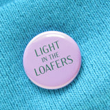 A round lavender pinback button that reads LIGHT IN THE LOAFERS in forest green. Button is on a aqua knit sweater.