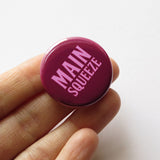Round magenta-maroon pinback button that reads MAIN SQUEEZE in a light pink font. Button is held in a white hand.