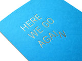 Close-up photo of a  bright medium blue greeting card with off white envelope, reads HERE WE GO AGAIN in a thin metallic silver foil, sans serif font. The word AGAIN starts to slide off, creating the image of the word coming apart and falling downward