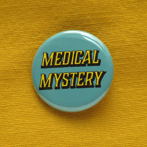 Round pinback button that says MEDICAL MYSTERY. Yellow shadowed text on a scrubs blue background. Button is pinned to a mustard yellow fabric.