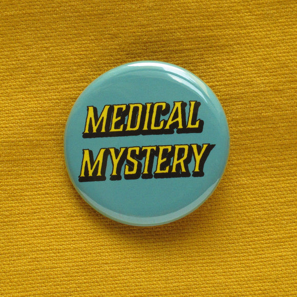 Round pinback button that says MEDICAL MYSTERY. Yellow shadowed text on a scrubs blue background. Button is pinned to a mustard yellow fabric.