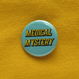 Round pinback button that says MEDICAL MYSTERY. Yellow shadowed text on a scrubs blue background. Button is pinned to a mustard yellow fabric.