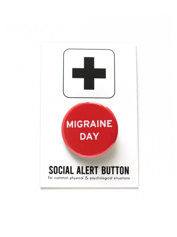 Shiny bright red pinback button with white san serif test reading, Migraine Day.  The button is attached to a white backing card with a black plus sign at the top of the card, and beneath the button it reads: Social Alert Button, for common physical & psychological situations.