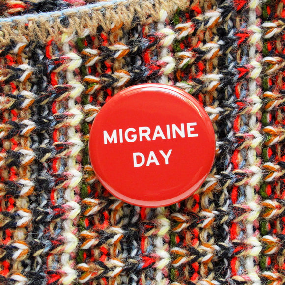 Shiny bright red pinback button with white san serif test reading, Migraine Day.  The button is pinned to a multicolor knit sweater with flecks of cream, black, brown read and pale blue.