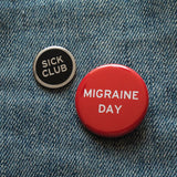 Round enamel pin that says SICK CLUB next to a red pinback button that says MIGRAINE DAY. 