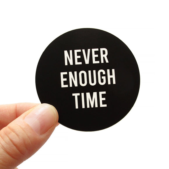 A round black sticker that reads NEVER ENOUGH TIME in white san serif text.  The sticker is being held in the lower left hand corner by a white thumb and forefinger.