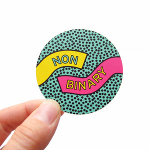 Multicolor circular stickers held at the left corner with a white thumb and forefinger. Sticker backgrounder is teal with tiny black dots. A ribbon of yellow comes from the left side and reads NON and a ribbon of pink comes in on the right side and reads BINARY. Sticker has a retro eighties Memphis design style. 