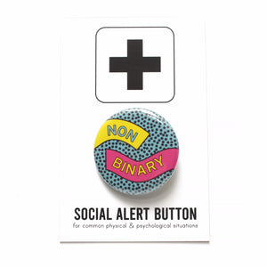Round pinback button reading NON-BINARY.  The buttons has a  teal background and tiny black dots, a yellow ribbon comes in on the left reading NON, and a pink ribbon on the right comes in reading BINARY. The button is on a white card with a black plus sign at the top, at the bottom is reads Social Alert Button for common physical, psychological & social situations