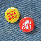 Two shiny pinback buttons on a denim background.  The button on the left is bright yellow with red-orange san serif text that reads OVER WORKED. The pinback button on the right is vibrant red-orange and reads UNDER PAIN in white san serif text.