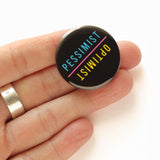 Pinback button that says Pessimist in one direction, and Optimist in the opposite direction.  The button sits in an open hand for size reference.