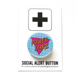 Round light pinback button with a lavender grid pattern, and magenta pink triangle reads POWER DYKE in golden yellow text. On a white backing card, black plus sign at the top. Below reads: Social Alert Button, with tiny text beneath it which says, for common physical & psychological situations