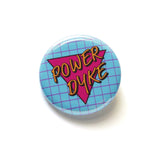 Round light pinback button with a lavender grid pattern, and magenta pink triangle reads POWER DYKE in golden yellow text.