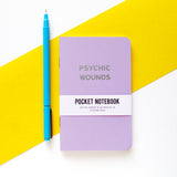 Lavender pocket notebook that says PSYCHIC WOUNDS in silver text, next to a pen for size reference.  The height of the notebook is about the same as the pen.