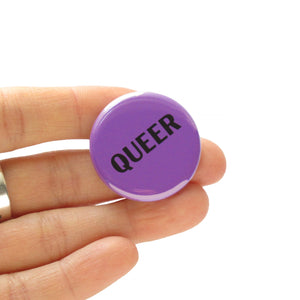 Round purple pinback button that reads QUEER in black text. Held in a hand.