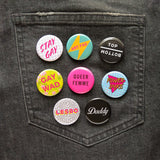 Black jeans back pocket with a bunch of bright and shiny gay & queer pinback buttons pinned on. Featuring Stay Gay, Art Fag, Top/Bottom, Gay Wad, Queer Femme, Power Dyke, Less, Daddy