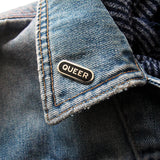 Capsule shaped silver pin with black enamel that reads QUEER. Pin is on the lapel of a faded blue denim jacket.