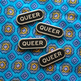 Four capsule shaped pins that say QUEER on a blue patterned fabric background.