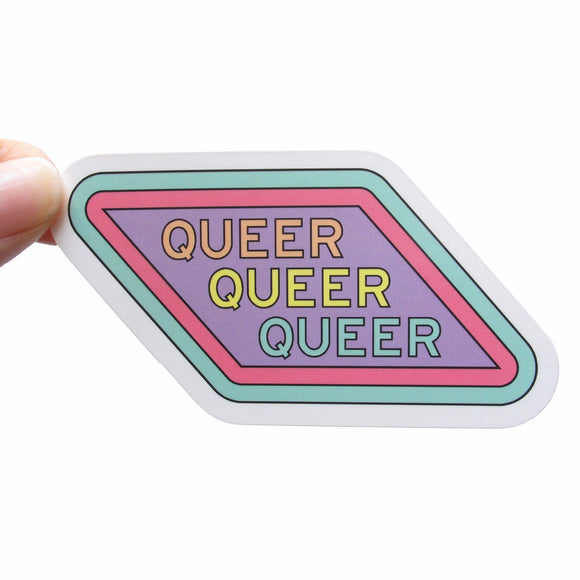 Parallelogram sticker that reads QUEER three times, ones in peach, once in yellow and lastly in pastel teal. The sticker background is lavender with a pink outline and a pastel teal outline. The sticker is held by a white thumb and forefinger in the upper left corner.