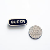Capsule shaped enamel pin that says QUEER next to a dime for size reference.