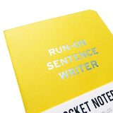 Close-up of a bright yellow small notebook with rounded corners, and a blue-silver imprint reading RUN-ON SENTENCE WRITER