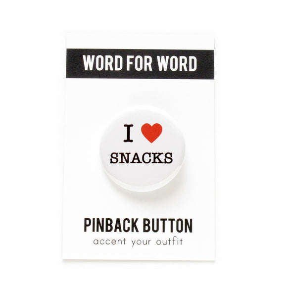 A round white button that reads I LOVE SNACKS. Love being represented by a red heart. Button is pinned to a Word For Word branded backing card.