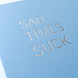 Powder blue greeting card that says SAD TIMES SUCK. Close up of the silver hot foil pressed text.