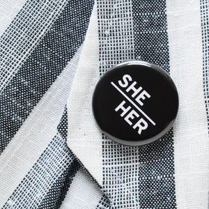 Round pinback button with black background which reads SHE/HER on an angle in white text, in a san serif font.  Button is pinned on a white & gray textile blazer lapel.