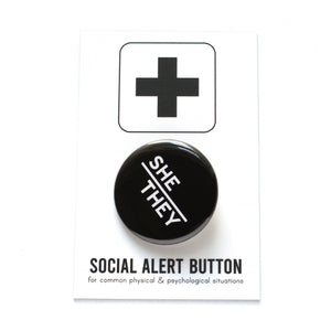 Round pinback button with black background which reads SHE/THEY  on an angle in white text, in a san serif font. On a white backing card, black plus sign at the top. Below reads: Social Alert Button, with tiny text beneath it which says, for common physical & psychological situations