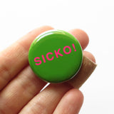 Round rich green pinback button reads SICKO! in hot pink text. On a white backing card, black plus sign at the top. Held in a white hand with band-aid on one finger
