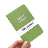 Hand holding up a moss green pocket notebook that says SMART STONER on the cover.