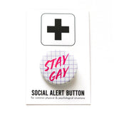 Round white pinback button with a lavender grid pattern reads STAY GAY in hot pink text. On a white backing card, black plus sign at the top. Below reads: Social Alert Button, with tiny text beneath it which says, for common physical & psychological situations
