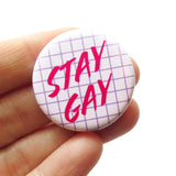 Round white pinback button with a lavender grid pattern reads STAY GAY in hot pink text. Button is being held by a white hand.