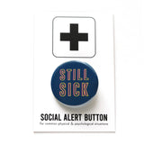 Dark blue round pinback button with neon yellow & pink text that reads STILL SICK. Button is on a Social Alert Button backing card.