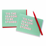 Two light green holiday Christmas cards with white text and a red drop shadow, that read ’Tis The Season To Be Stoned. They have red envelopes, and one is at an angle with a red pen across the upper corner