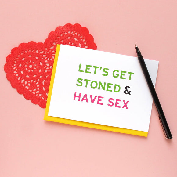 Light pink background with red paper lace heart, and a greeting card that says Let's Get Stoned & Have Sex in green and pink san serif text.  A black Le Pen lays across the edge of the card.