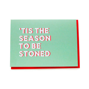 Two light green holiday Christmas cards with white text and a red drop shadow, that read ’Tis The Season To Be Stoned. They have red envelopes, and one is at an angle with a red pen across the upper corner