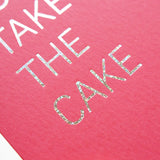 Close up of TAKE THE CAKE greeting card with the silver glitter embossed word of CAKE in sharp focus in the foreground
