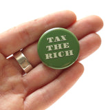 TAX THE RICH, pinback button sitting in an open hand, for size reference.